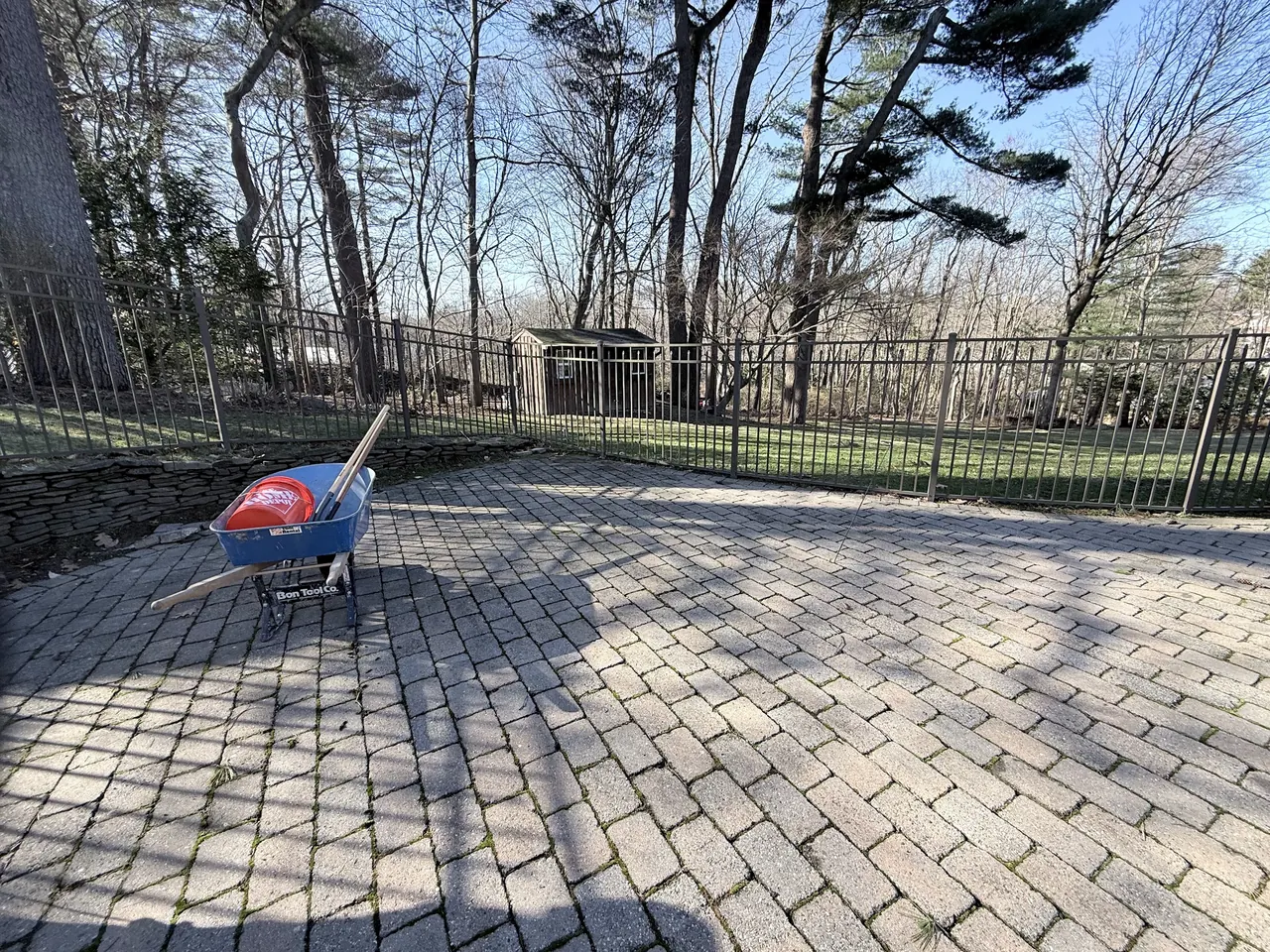 Existing backyard paver patio before gazebo installation, with a fenced lawn and trees