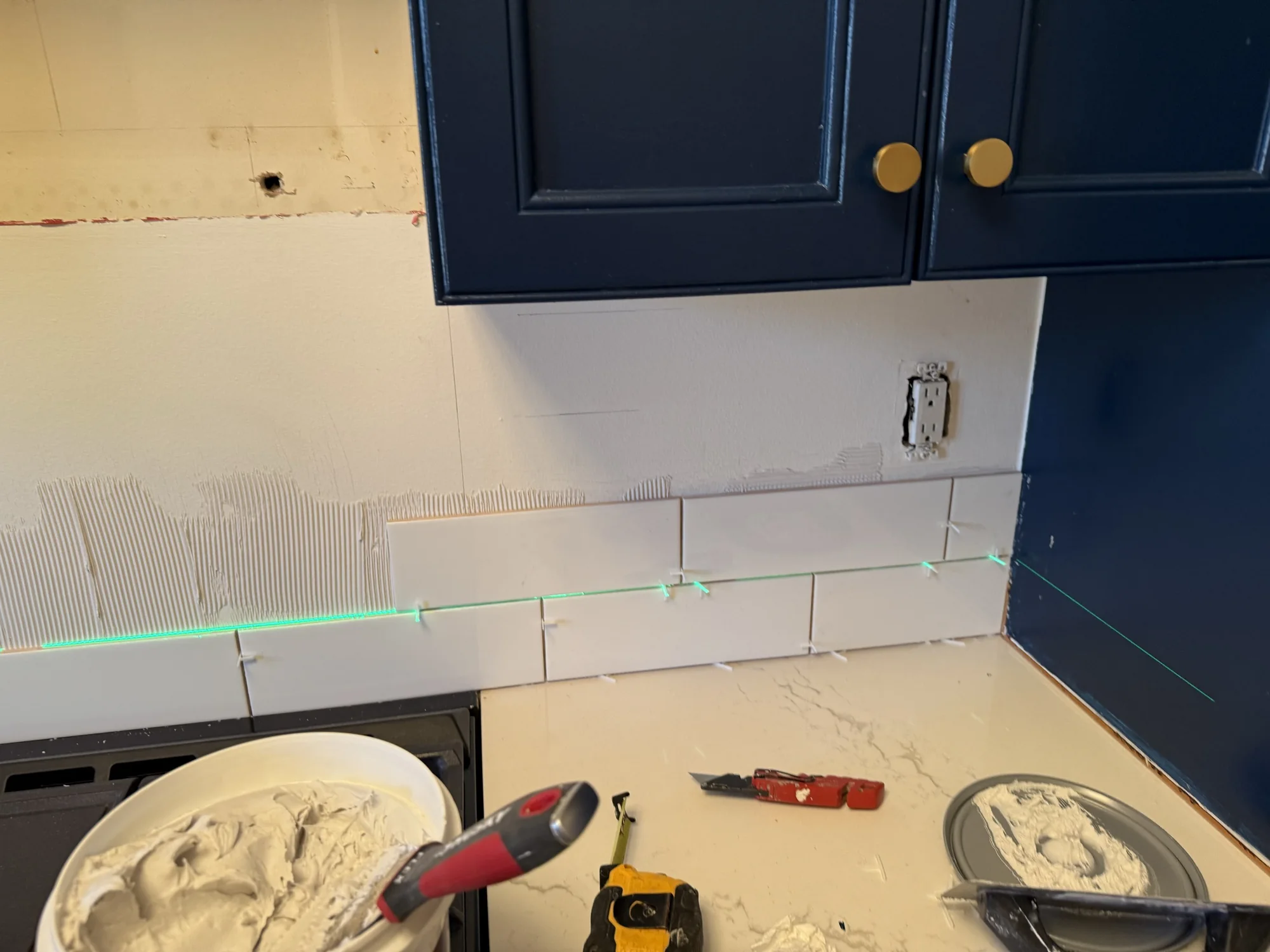 First rows of white subway tile set along a green laser line above the countertop, with a tub of mortar and tools