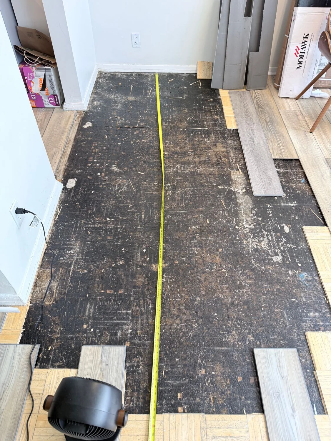 Tape measure across the bare subfloor to size the plywood replacement, Bronx apartment