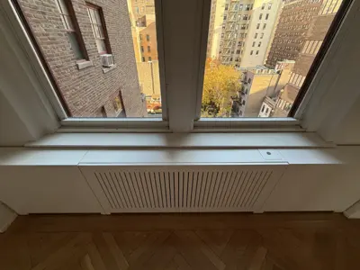 Newly installed white radiator cover beneath a window with NYC cityscape view and herringbone hardwood floor